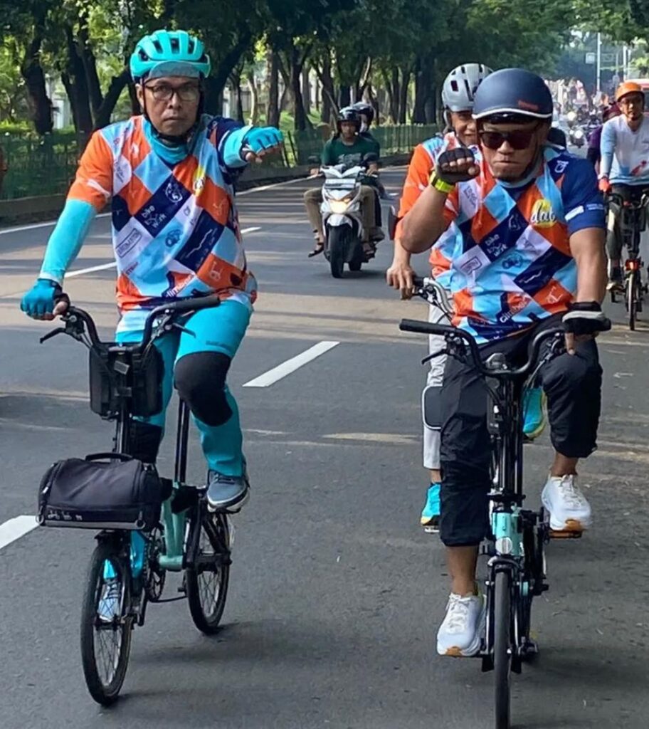 Group of cyclists riding on a city street wearing matching orange blue geometric jerseys and teal helmets loweringor raising fists as they ride